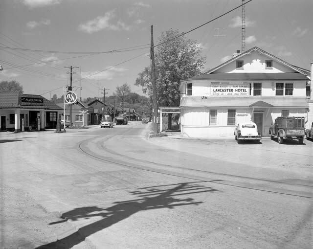 A historic black‑and‑white photograph of the Lancaster Hotel at a mid‑century Bridgeport intersection, showing the hotel’s signage, parked vehicles, and neighbouring gas station; this original print hangs inside The Lancaster Smokehouse as part of its heritage collection.
